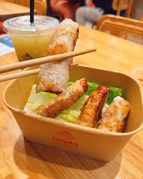 Chopsticks lifting a crispy spring roll over lettuce in a Nón Lá Express box